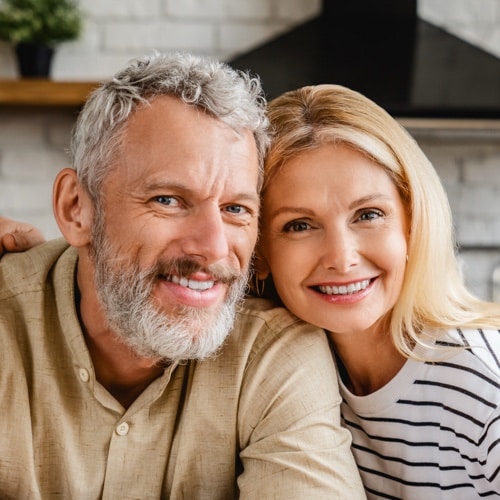 Pareja de mediana edad sonriendo, sentada en una cocina luminosa y mirando a la c&aacute;mara
